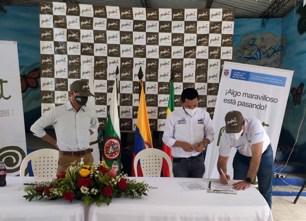 Tres hombres del equipo de la ANT en mesa de conversación, cada uno con tapabocas y camisa blanca, uno de ellos firmando un documento sobre la mesa. Encima de la mesa un arreglo de flores.