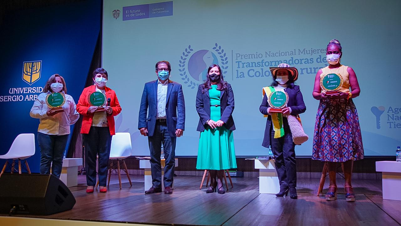 Fotografía de la celebración y entrega del premio nacional MUJERES TRANSFORMADORAS DE LA COLOMBIA RURAL. Sobre una tarima con piso de madera y de fondo el logo de la premiación, están cuatro mujeres ganadoras de este premio en compañía de la Directora de la ANT y un funcionario hombre de la ANT. 
