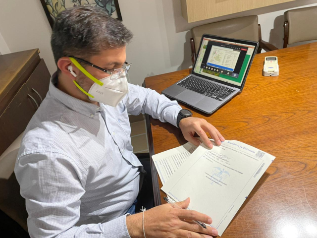 Hombre lleva puesto camisa blanca, tapabocas blanco. Sobre mesa de escritorio él apoya los brazos, así como un computador portátil. En sus manos tiene algunas hojas de títulos de propiedad.