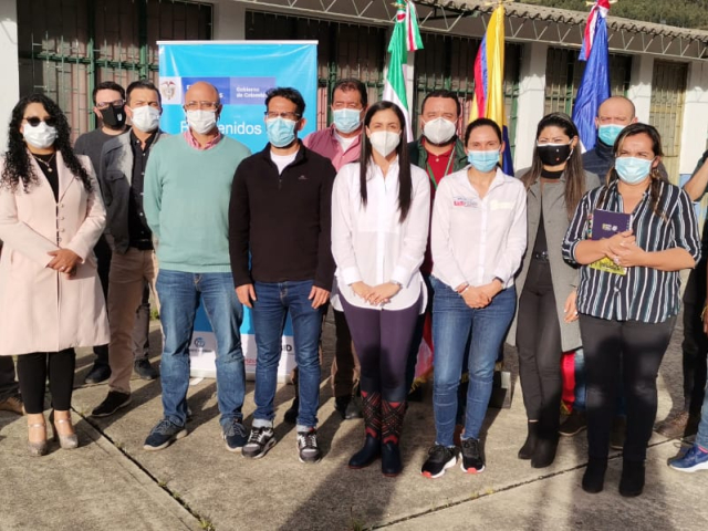 Diez hombres y siete mujeres posan de pie para la toma de una fotografía. De fondo una vaya vertical con el logo del Gobierno, y tres banderas entre ellas la de Colombia. 