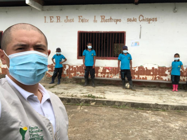 Selfi tomada por un hombre funcionario de la ANT. Él de medio cuerpo, luce el chaleco corporativo de la ANT y lleva puesto camisa blanca y tapabocas. De fondo aparece la institución educativa José Félix Restrepo sede Chispas y cuatro niños estudiantes vistiendo todos camisas iguales de color azul, pantalón negro y botas de caucho. 