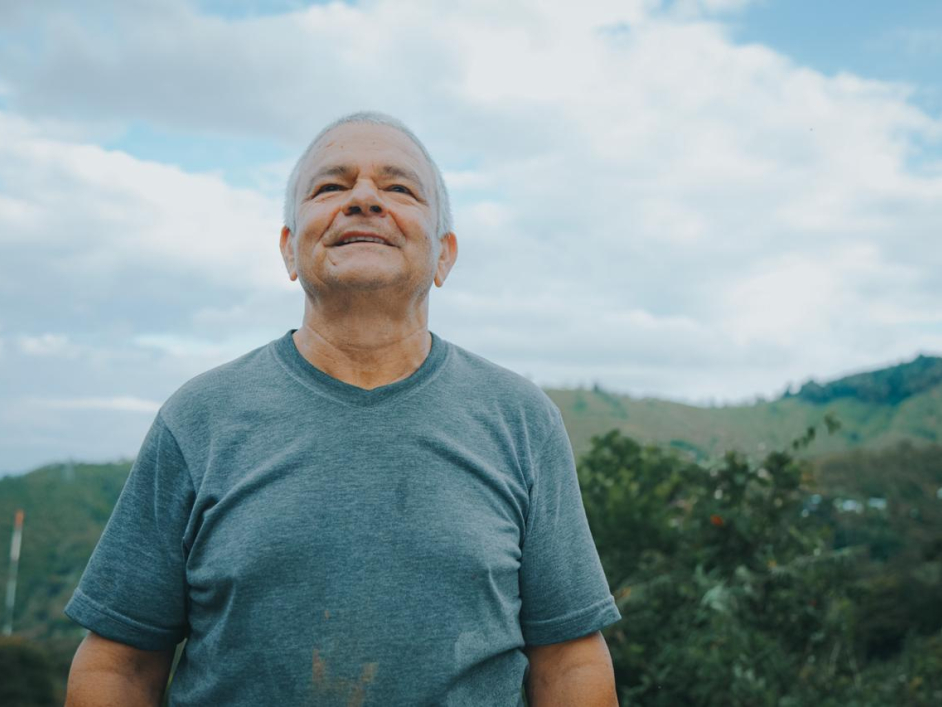 Aparece un señor, adulto mayor, de medio cuerpo, al aire libre, sonriente, con una camiseta color azul y de fondo las montañas verdes.