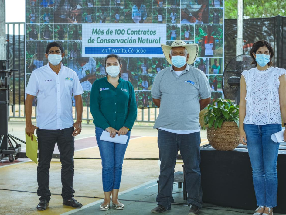 La Directora de la ANT, Myriam Martínez Cárdenas, aparece de pie y cubriendo sus bocas con tapabocas, con tres personas más, campesinos. De fondo una pancarta con varias fotografías de campesinos beneficiados de la entrega de títulos.