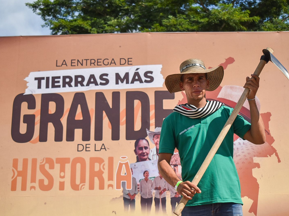 Campesino con su azadón y un cartel detrás que dice "La entrega de tierras más grande de la historia".